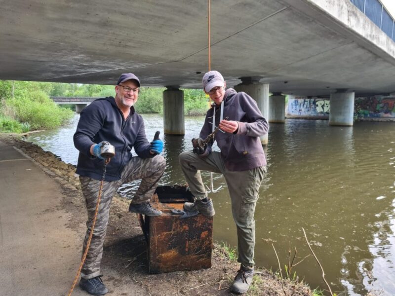 Twee mannen poseren onder een brug tijdens het magneetvissen en tonen hun vondst: een grote, roestige kluis uit het water.