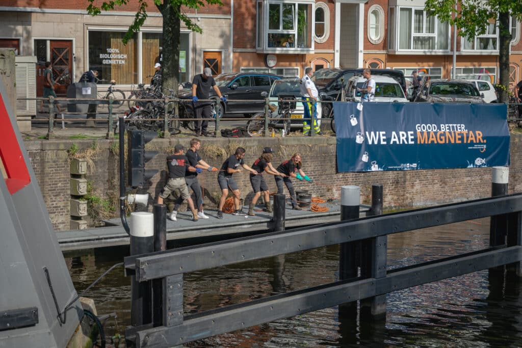 In een stad zijn 5 mensen erg hard aan een touw aan het trekken tijdens het magneetvissen. Naast de mensen staat een spandoek 'we are magnetar'
