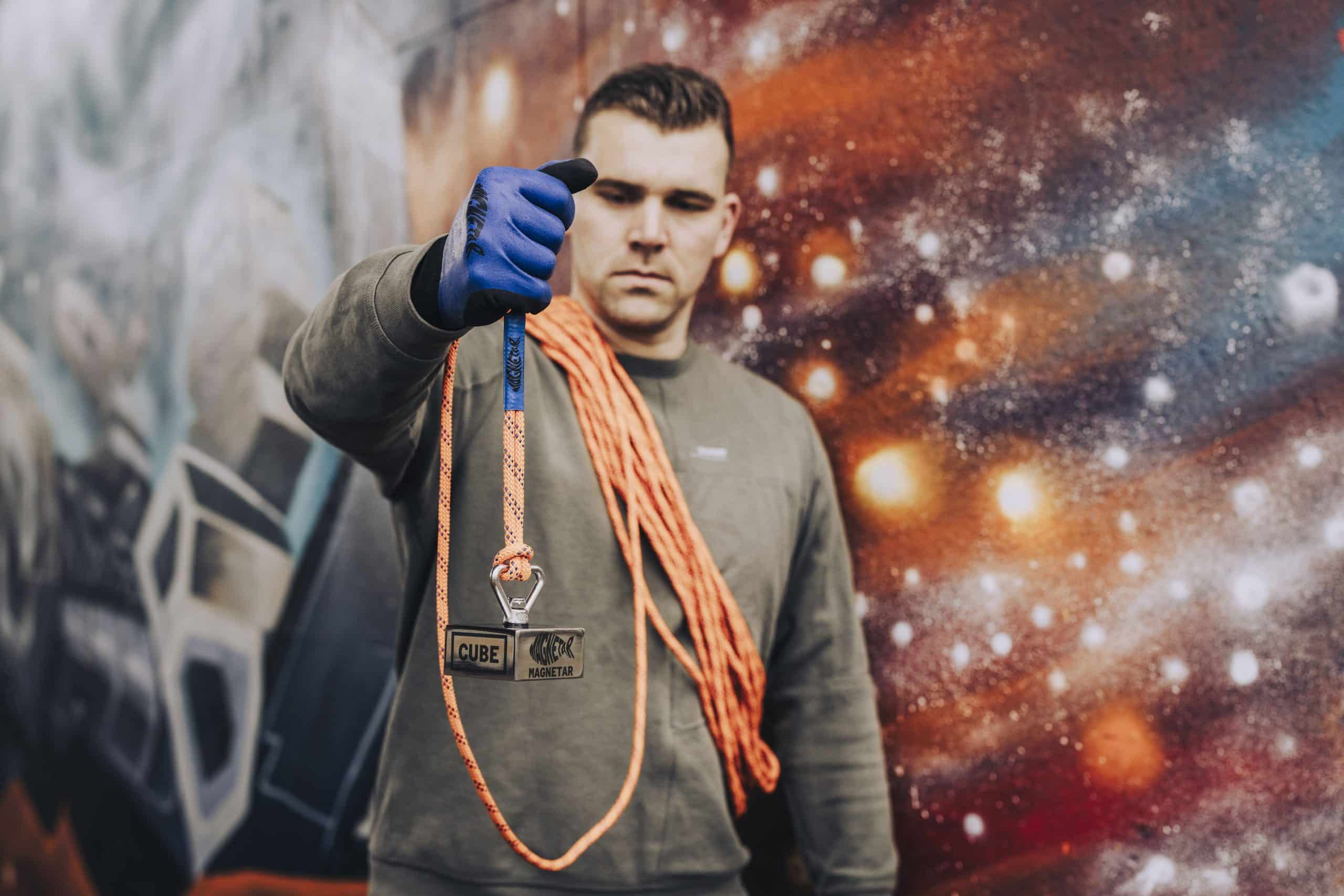Een man houdt de Magnetar Cube magneet vast aan een oranje touw, met de schouder vol touw en een kleurrijke kosmische graffiti-achtergrond.