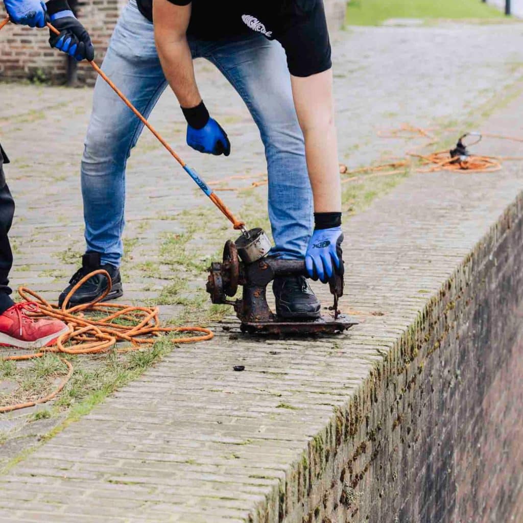 2 mannen trekken aan een touw met daaraan een magneet. De magneet kleeft vast aan een oude roestige naaimachine die ze uit het water getrokken hebben tijdens het magneetvissen